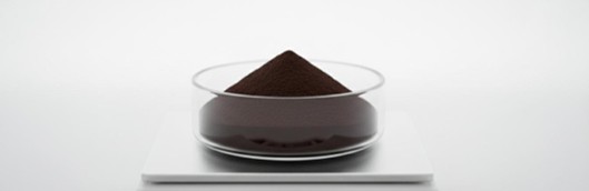 Dark brown iron oxide magnetic nanoparticle powder in a glass weighing dish on an analytical balance.