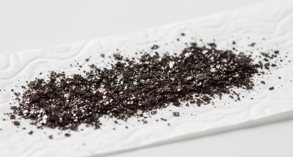 Dark brown-black graphene oxide powder with flaky layered morphology spread on white weighing paper.