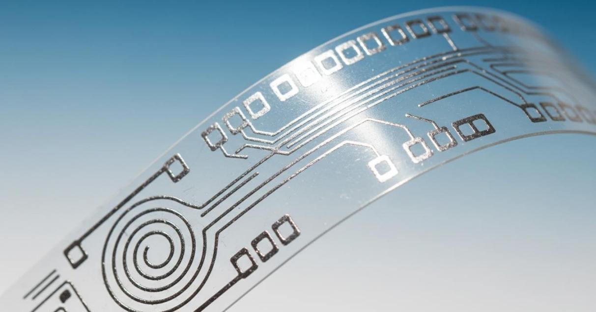 Flexible printed circuit with silver conductive traces and antenna pattern on transparent PET substrate demonstrating bendability